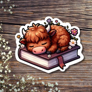 May include: A brown and white Highland cow sleeps on a closed book with a brown cover. The book is surrounded by pink, white and purple flowers.