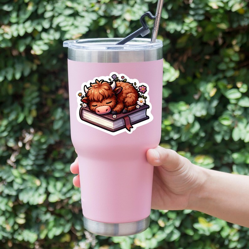 May include: A pink stainless steel tumbler with a sticker of a brown highland cow sleeping on a book with flowers around it.