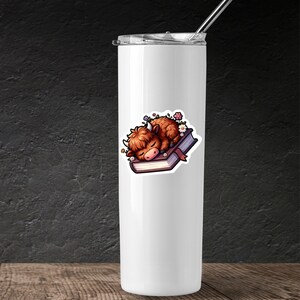 May include: A white stainless steel tumbler with a sticker of a brown highland cow sleeping on a closed book with flowers around it.