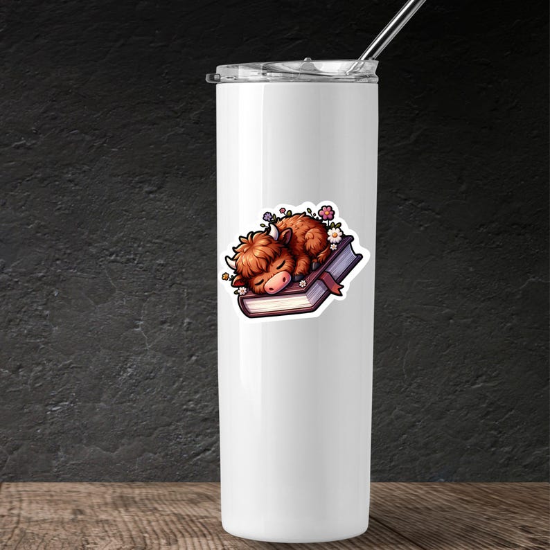 May include: A white stainless steel tumbler with a sticker of a brown highland cow sleeping on a closed book with flowers around it.