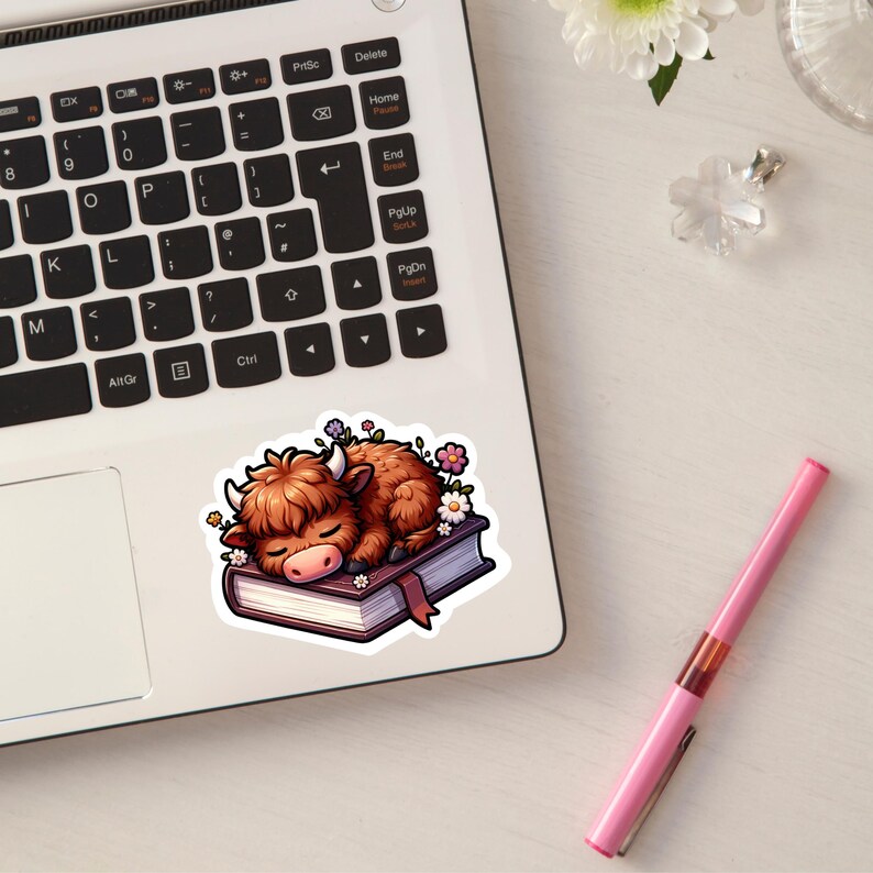 May include: A brown and white Highland cow with a fluffy mane is asleep on a closed book with a brown leather cover. The book is surrounded by pink and white flowers.