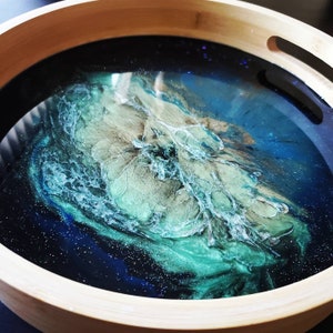 May include: A round wooden serving tray with a resin design. The resin features shades of blue, teal, and gold, creating a swirling, abstract pattern. The tray has a natural wood rim and integrated handles.