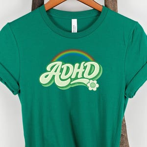 May include: A green t-shirt with rolled-up sleeves. The shirt features the word "ADHD" in a retro font with a rainbow and flower design. The shirt is hanging on a wooden hanger against a white background.