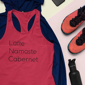 Pu&ograve; includere: Un top rosso senza maniche con il testo "Latte Namaste Cabernet" e la parola "HEALTHAHOLIC" in basso. Il top &egrave; sovrapposto a una giacca blu. L'immagine include anche una bottiglia d'acqua nera, un paio di scarpe da corsa arancioni e nere, uno smartphone nero e auricolari bianchi.