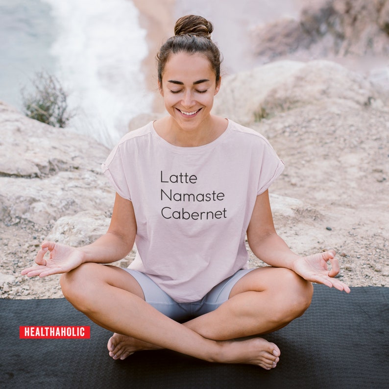 Pu&ograve; includere: Una donna indossa una maglietta rosa con la scritta "Latte Namaste Cabernet" e siede in una posa yoga su un tappetino da yoga nero.
