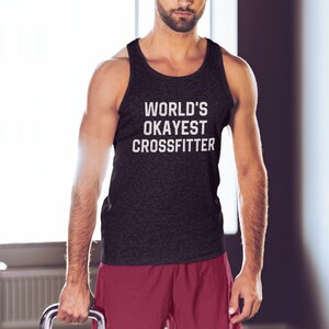 May include: A black tank top with the text "WORLD'S OKAYEST CROSSFITTER" in white. A man is wearing the tank top and holding a black kettlebell with a silver handle. The kettlebell has the number 20 printed on it. The man is wearing red athletic shorts.
