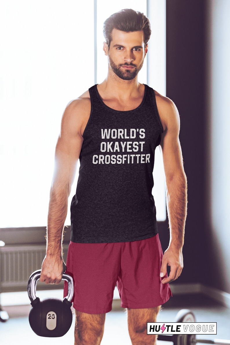 May include: A black tank top with the text "WORLD'S OKAYEST CROSSFITTER" in white. A man is wearing the tank top and holding a black kettlebell with a silver handle. The kettlebell has the number 20 printed on it. The man is wearing red athletic shorts.