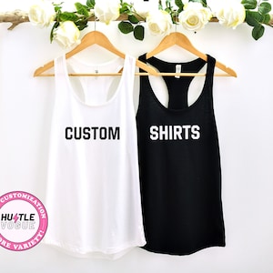 May include: Two tank tops hanging on wooden hangers. The white tank top says "CUSTOM" in black letters. The black tank top says "SHIRTS" in white letters.