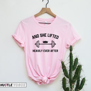 May include: Pink t-shirt with a black graphic that says "And She Lifted Heavily Ever After" with a crown and barbell.