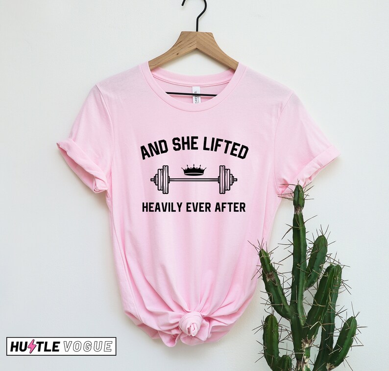 May include: Pink t-shirt with a black graphic that says "And She Lifted Heavily Ever After" with a crown and barbell.