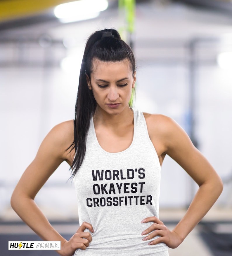 May include: A woman wearing a gray tank top with the text "WORLD'S OKAYEST CROSSFITTER" printed on it. She is standing in a gym setting.