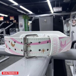 Peut inclure: Ceinture de musculation blanche avec un motif rayé rose et blanc. La ceinture a un motif d'étoile dorée et de croissant de lune et le texte "EYSM" imprimé sur le côté.