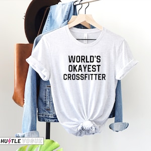 May include: White t-shirt with black text that reads "WORLD'S OKAYEST CROSSFITTER".
