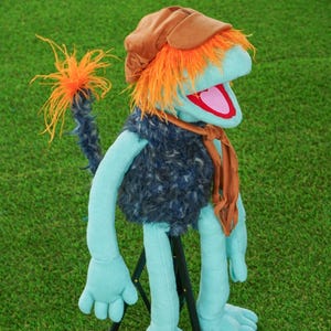 May include: A light blue puppet with bright orange hair, wearing a brown cap, a dark blue fuzzy vest, and a brown scarf. The puppet has a long tail with orange tufts at the end, a wide open mouth with a pink interior, and is supported by a black stand.
