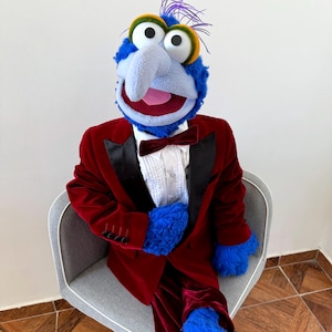 May include: A blue puppet with large eyes, a long nose, and purple hair, wearing a red velvet tuxedo with black lapels and a matching bow tie. The puppet is seated in a grey chair.