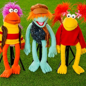 May include: Three colorful puppets with distinct features. The left puppet has purple hair, an orange face, and a yellow and red striped shirt. The center puppet has orange hair, a brown cap, and a blue body. The right puppet has red hair and a red shirt.