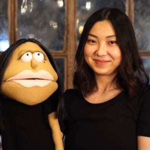 May include: A woman with long dark hair is smiling and holding a puppet. The puppet is a woman with long dark hair and a neutral expression. The puppet is wearing a black shirt.