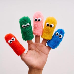 May include: A hand displaying five colorful finger puppets. Each puppet is a different color: red, green, pink, yellow, and blue. They have fuzzy bodies and large googly eyes, creating a playful and whimsical appearance.