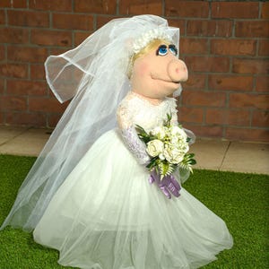 May include: A pig puppet in a white wedding dress, veil, and floral headpiece. The puppet holds a bouquet of white flowers and wears purple gloves. The background includes a brick wall and a green surface.