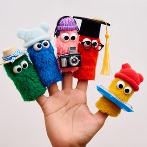 May include: A hand displaying five finger puppets, each with large googly eyes and unique accessories. The puppets are various colors, including green, blue, pink, red, and yellow. Accessories include a hat, headphones, a camera, glasses, and a graduation cap.