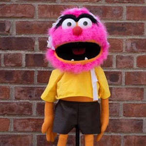 May include: A pink furry puppet with a yellow shirt, black shorts, and a white headband. The puppet has a large mouth and black eyes.