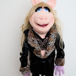 Peut inclure: Marionnette de cochon en peluche avec des oreilles roses, un grand museau et des yeux bleus. La marionnette porte une veste noire avec des bordures léopard, un haut en dentelle noire, un collier de perles et des gants lavande avec une bague.
