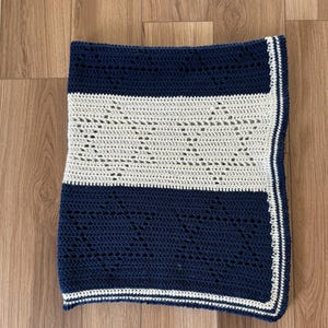 Op de afbeelding: Een gehaakt babydekentje met een gestreept patroon in marineblauw en wit. Het dekentje heeft een witte rand en is gemaakt met een delicate steek.