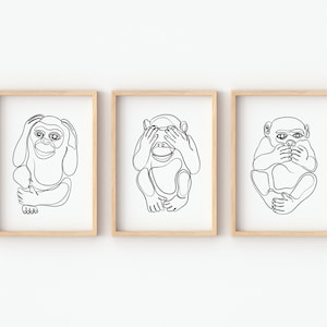 Op de afbeelding: Set van drie ingelijste kunstprints met minimalistische lijntekeningen van apen. Elke aap wordt in een andere pose afgebeeld: oren, ogen en mond bedekkend. Het kunstwerk is in zwart op een witte achtergrond, ingelijst in licht hout.