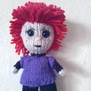 May include: A knitted doll with red hair, a purple shirt, and black pants. The doll has a white face with blue eyes and a knitted frown.