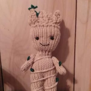 May include: A knitted plush toy of the character Groot from the Guardians of the Galaxy franchise. The toy is beige with green leaves and has a smiling face.