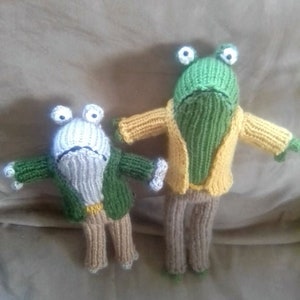 May include: Two knitted frog plushes wearing green jackets and brown pants. The larger frog is green with white eyes, and the smaller frog is white with black eyes.