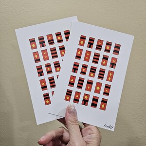 May include: Two white cards with a repeating pattern of red and black rectangles with yellow, red, and black geometric shapes inside. The cards are stacked on top of each other and the bottom card has the word "keofelt" printed in black at the bottom right corner.