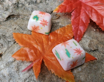 May include: Two white ceramic keycaps with a pink and green floral design. The keycaps are on top of a bright orange maple leaf.