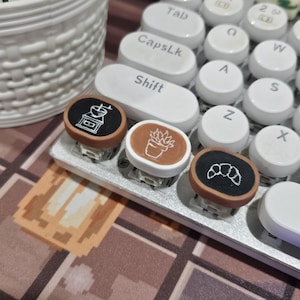 May include: Three round, brown and white keycaps with different designs. The keycaps feature a coffee grinder, a potted plant, and a croissant.