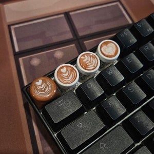 May include: Four brown and white ceramic keycaps with latte art designs on a black keyboard. The keycaps feature different latte art designs, including a heart, a leaf, and a swirl.