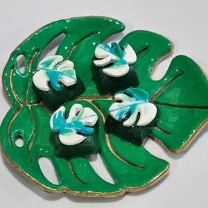 May include: Four green and white resin keycaps with a monstera leaf design. The keycaps are sitting on a green leaf-shaped dish.