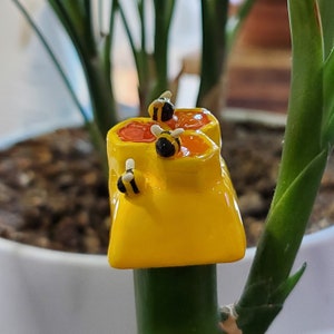 May include: A yellow ceramic honey pot with three small bees perched on top. The honey pot has a red and orange glaze and is decorated with white accents.