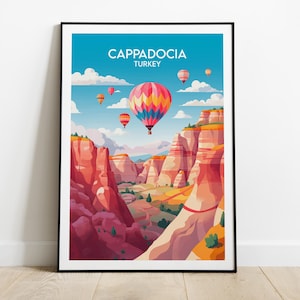 May include: A framed art print showing a vibrant illustration of Cappadocia, Turkey. The image features hot air balloons above a canyon with red rock formations. The sky is blue with clouds, and the print is in a black frame.