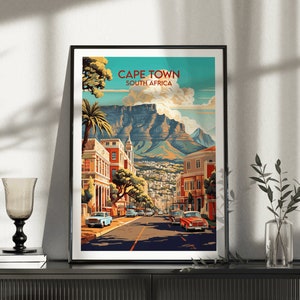 May include: A vintage-style travel poster featuring a colorful illustration of Cape Town, South Africa. The poster depicts a cityscape with buildings, palm trees, and a mountain in the background. The text "CAPE TOWN SOUTH AFRICA" is displayed at the top of the poster.