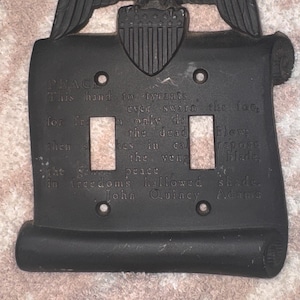 Puede incluir: Cubierta de interruptor de luz de metal negro con un águila y un escudo, y un diseño de pergamino. Tiene dos recortes para interruptores y texto grabado de John Quincy Adams. Medidas en centímetros.