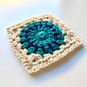 May include: A crocheted square with a teal and purple center surrounded by a cream border.