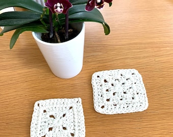 May include: Two white crocheted square coasters on a wooden surface. A potted plant with purple orchids is in the background.