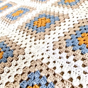 Pode incluir: Um cobertor de crochê com um padrão repetitivo de quadrados em branco, bege, azul e amarelo.