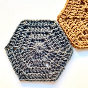 May include: Two crocheted hexagons, one in a dark grey colour and one in a light brown colour. The hexagons have a lacy pattern and are made with a thick yarn.