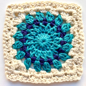 Op de afbeelding: Een gehaakt vierkant met een blauw en paars bloemenontwerp in het midden, omringd door een witte rand.