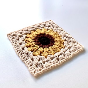 Puede incluir: Un cuadrado tejido a crochet con un diseño de girasol en el centro. El girasol está hecho con hilo marrón, amarillo y negro. El cuadrado está hecho con hilo beige.