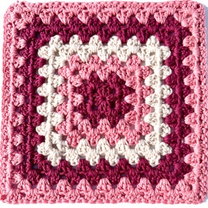 Può includere: Un quadrato di granny crochet con un design multicolore. Il centro del quadrato è di colore bordeaux scuro, circondato da un anello rosa chiaro, poi da un anello bianco e infine da un anello bordeaux. Il bordo esterno del quadrato è di colore rosa chiaro.