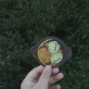 May include: A sticker depicting a dish with two white, shell-shaped dumplings and a brown sauce. The sticker has a dark brown border. The image is held against a backdrop of green foliage.