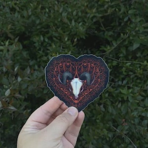 Puede incluir: Una pegatina en forma de corazón con un diseño de calavera de carnero, con cuernos curvos y un patrón ornamental rojo y negro. La pegatina se sostiene en una mano, con un fondo de follaje verde.