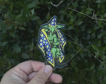 May include: A sticker featuring a green goblin wizard wearing a blue and yellow star-patterned robe and hat. The goblin is holding a smaller goblin. The sticker is held in front of a background of green leaves.